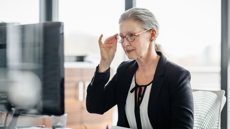 Woman grapping her glasses looking at a desktop computer